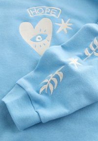 Felpa azzurro chiaro con grafiche ricamate bianche: un cuore, un occhio, stelle e la parola "SPERANZA." Texture morbida con polsini a costine.