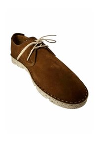 Zapato de ante marrón con forma redondeada, cordones beige y suela blanca moteada. Presenta costuras en contraste alrededor del borde.
