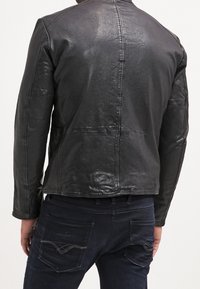 Uomo che indossa una giacca in pelle nera attillata e jeans blu scuro, mostrato di spalle su uno sfondo semplice.