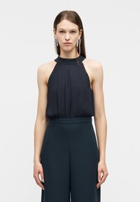 Γυναίκα που φοράει μαύρο αμάνικο τοπ με ψηλό λαιμό και μαύρο παντελόνι με φαρδιά πόδια, με αξεσουάρ μακριά κρεμαστά σκουλαρίκια με κρυστάλλους, σε απλό φόντο.