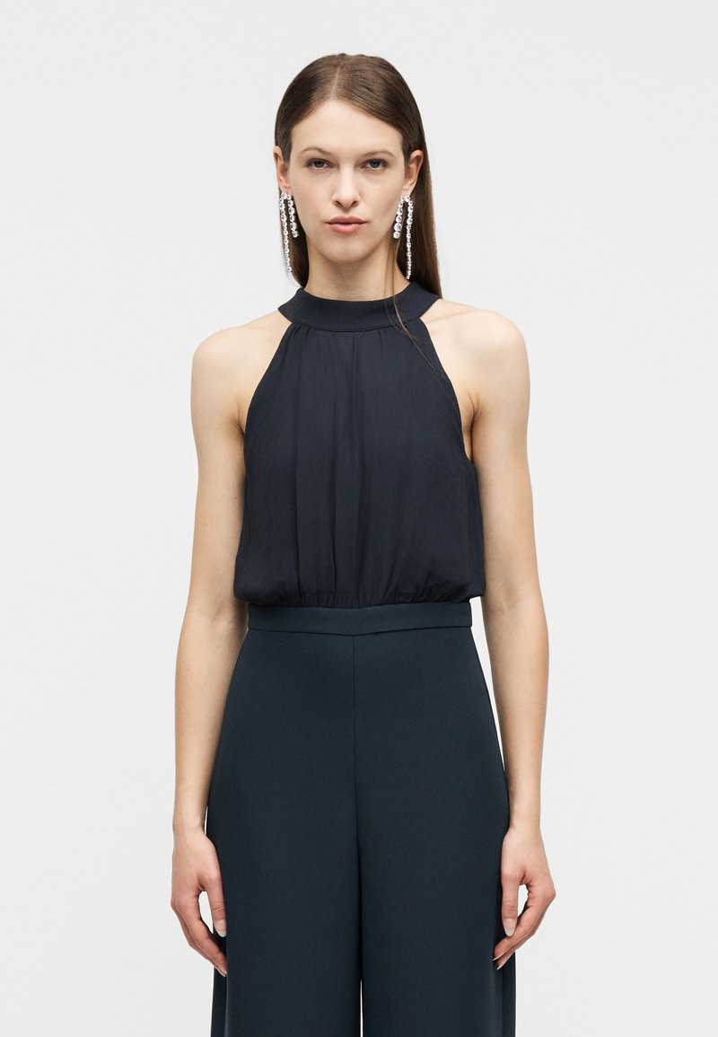 Γυναίκα που φοράει μαύρο αμάνικο τοπ με ψηλό λαιμό και μαύρο παντελόνι με φαρδιά πόδια, με αξεσουάρ μακριά κρεμαστά σκουλαρίκια με κρυστάλλους, σε απλό φόντο.