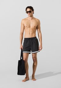 Fekete úszónadrág fehér díszítéssel, húzózsinóros derékkal és anyagtextúrával; fekete táskával és sötét napszemüveggel párosítva.