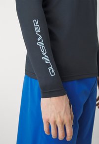 Čierna rash guard s dlhým rukávom, na ktorej je umiestnené logo Quiksilver svetlomodrej farby. Odev má hladkú textúru a priliehavý strih, ktorý je doplnený modrými šortkami.