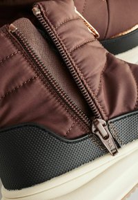 Stivale puffer marrone con parte superiore in tessuto trapuntato, caratterizzato da una zip prominente e suola nera testurizzata, accentuata da gomma beige.