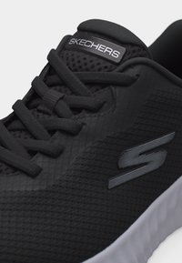 Μαύρο αθλητικό παπούτσι Skechers με επάνω μέρος από δίχτυ, μαύρα κορδόνια, ετικέτα με το λογότυπο στη γλώσσα και λευκή σόλα, εμφανές σε ανοιχτό φόντο.