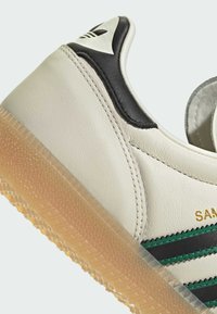 Krémové kožené tenisky s čiernymi a zelenými pruhmi, jantarovou gumovou podrážkou a polstrovaným čiernym pätkovým akcentom. Majú textúrovaný povrch a prešívané švy.