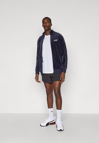 Tumesinine sametist zip-up jakki, millel on valge logo, koos valge T-särgi ja mustade lühikesed pükstega. Modell kannab valgeid ja musti sportjalatseid.