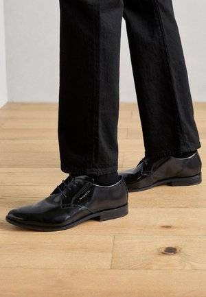 Personne portant des chaussures habillées noires et brillantes avec lacets et un pantalon noir, debout sur un sol en bois clair.
