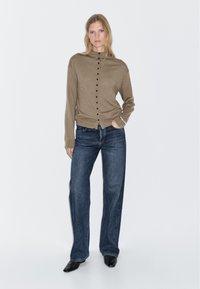 Beige stickad cardigan med hög hals och svarta knappar, i kombination med högmidjade, vida jeans i denim. Svarta skor fulländar looken.