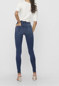 Mujer de espaldas, vestida con una blusa blanca de manga corta, jeans ajustados oscuros de tiro alto y sandalias transparentes de tacón alto, de pie contra un fondo liso.