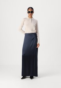 Lichtbeige lange mouwen polo shirt met knoopsluiting, gecombineerd met een marineblauw enkellang rokje. Model draagt grote zwarte zonnebril en oorbellen met een ring.