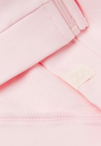 Světle růžová tkanina má měkkou strukturu, se zipovým zapínáním a tkanou etiketou s designem králíka. Jemné stehy jsou viditelné.