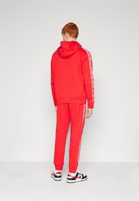 Rdeča kapuca in komplet trenirk s belimi stranskimi črtami. Narejeno iz mehke tkanine, z nastavljivo kapuco in rebrastimi zapestniki. Črne in bele superge.