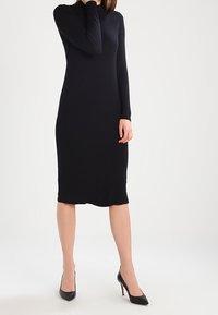 Schwarzes langärmliges Kleid in taillierter Form, knielang mit glatter Textur, kombiniert mit schwarzen high-heeled Schuhen.