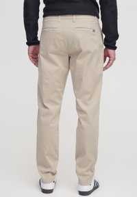 Beige cotton trousers shown from the back, featuring two rear pockets and a relaxed fit. Worn with white and black sneakers.
