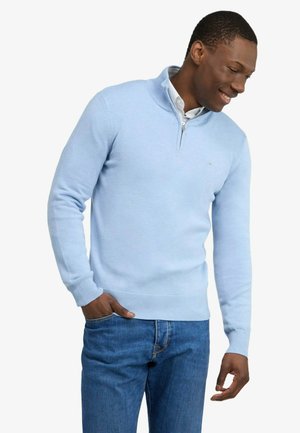 Homme portant un pull bleu clair à fermeture éclair quart, un jean bleu, souriant et regardant vers le bas à sa gauche.