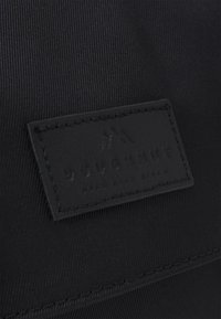 Zwarte stoffen tas met een gestructureerd oppervlak en een rechthoekige leren patch met een reliëflogo. Bevat stikdetails rond de patch.