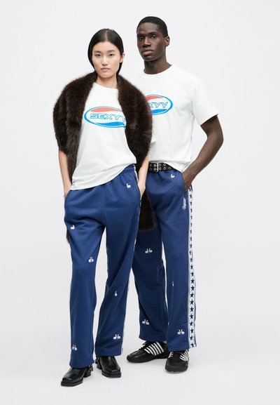 Vita grafiska t-shirts med texten "SEXYY", marinblå joggingbyxor med kanindekor och en brun pälsjacka. Kombination av enfärgade och mönstrade tyger.