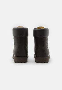 Botas de tobillo de cuero marrón con cuello acolchado, suela de goma resistente y detalle de logo en relieve en la parte posterior. Superficie lisa, diseño robusto.