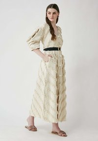 Maxime dress in beige with a textured zigzag pattern, puffed sleeves, and a black belt featuring a circular buckle. Pockets on both sides.