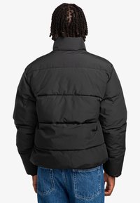 Sort dunjakke med høj krave, der har quiltede sektioner og en mat overflade. Kombineret med lyseblå denimjeans.