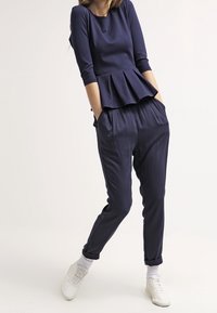 Kobieta ubrana w granatowy top peplum i dopasowane spodnie z tapered, stoi z rękami w kieszeniach i w białych sneakersach.