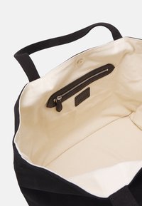 Black fabric tote bag with a beige canvas interior, featuring a zippered pocket, top handles, and a contrasting white lining.