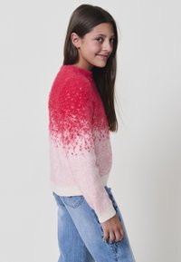 Fille aux longs cheveux noirs portant un pull fuzzy rose et blanc et un jean bleu clair avec broderies florales, souriant légèrement sur le côté.
