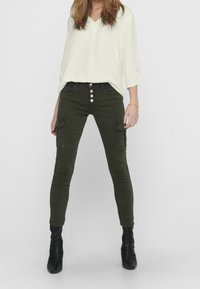 Pantaloni cargo verde oliva con vestibilità slim, cinque tasche e chiusura con bottone; abbinati a una blusa bianca a V e stivali neri con tacco.