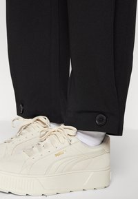 Black tailored pants feature a buttoned cuff, paired with beige sneakers, showcasing a textured surface and iconic branding.