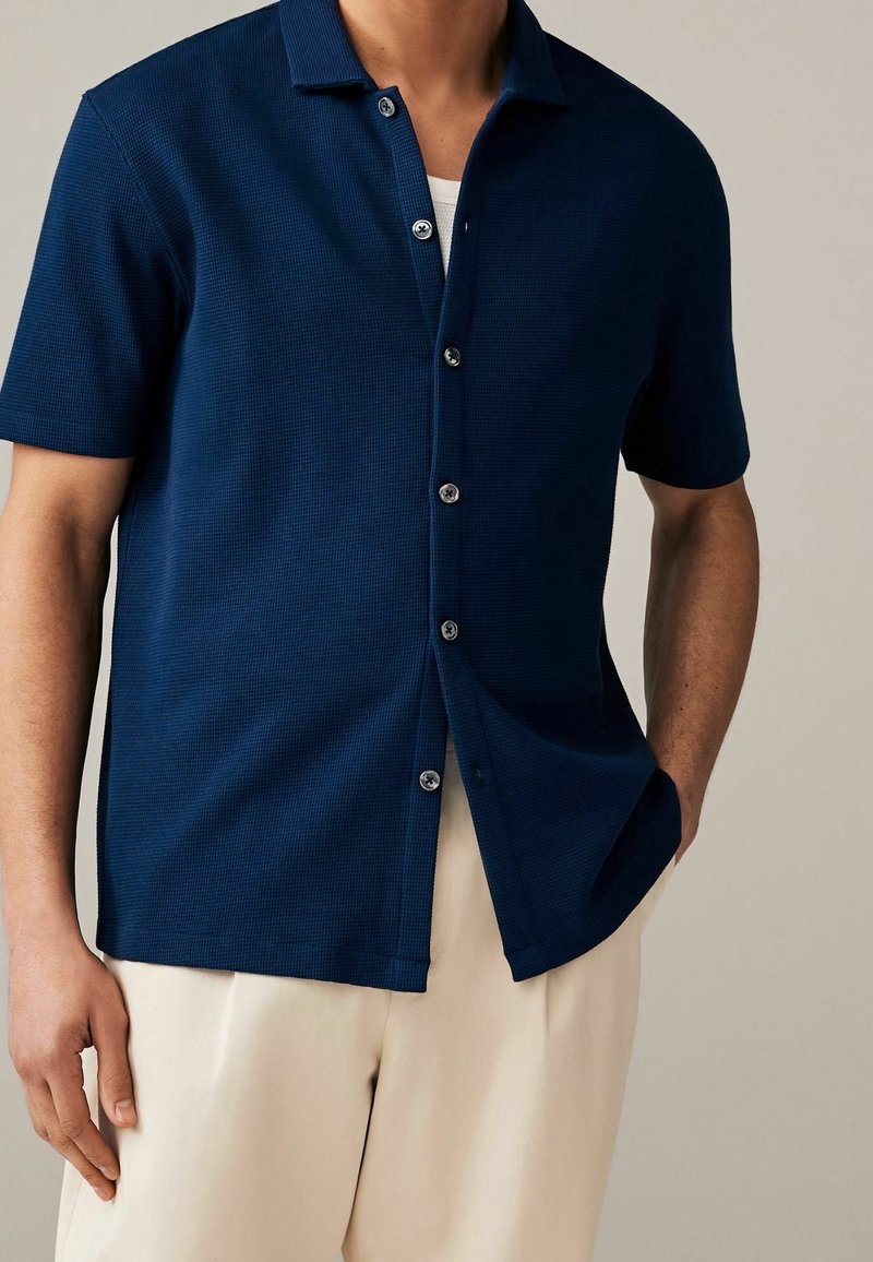 Chemise à manches courtes bleu marine à boutons, avec un motif texturé, un col et des boutons argentés, portée avec un pantalon de couleur claire.