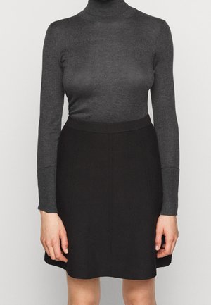 Femme portant un pull col roulé gris foncé ajusté et une jupe noire longueur genou, debout devant un fond clair uni.