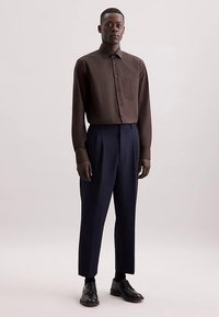 Camicia marrone a maniche lunghe con pattina di bottoni e tasca, abbinata a pantaloni sartoriali blu navy e scarpe nere, in piedi contro uno sfondo neutro.