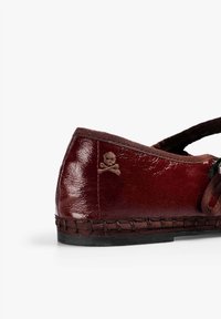 Zapato de cuero burdeos con punta redondeada, tira elástica y detalle bordado de calavera y tibias en el lateral, con bordes cosidos.
