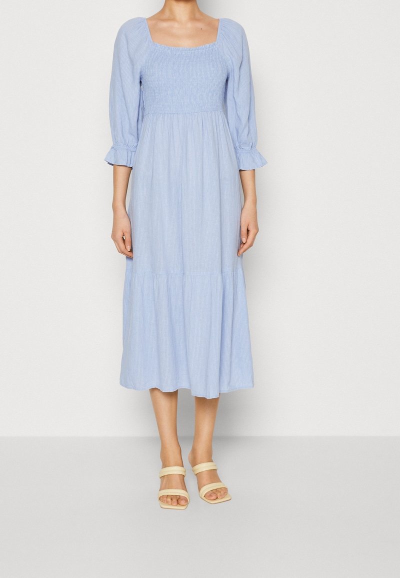 Femme portant une robe midi bleu clair avec un col carré froncé, des manches bouffantes 3/4, une jupe à volants et des sandales ouvertes beige.