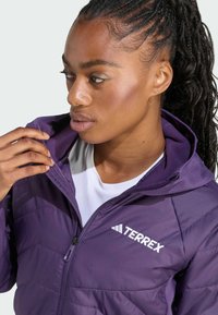 En ung kvinna med flätat hår bär en lila Terrex-jacka, där hon försiktigt drar i kragen och tittar eftertänksamt åt sidan.