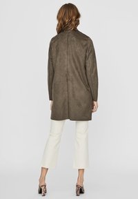 Oliivivihreä mokkavitja takki, jossa on suora malli, pitkät hihat ja takasauman yksityiskohta. Yhdistetty valkoisiin lyhyisiin housuihin ja korollisiin kenkiin.