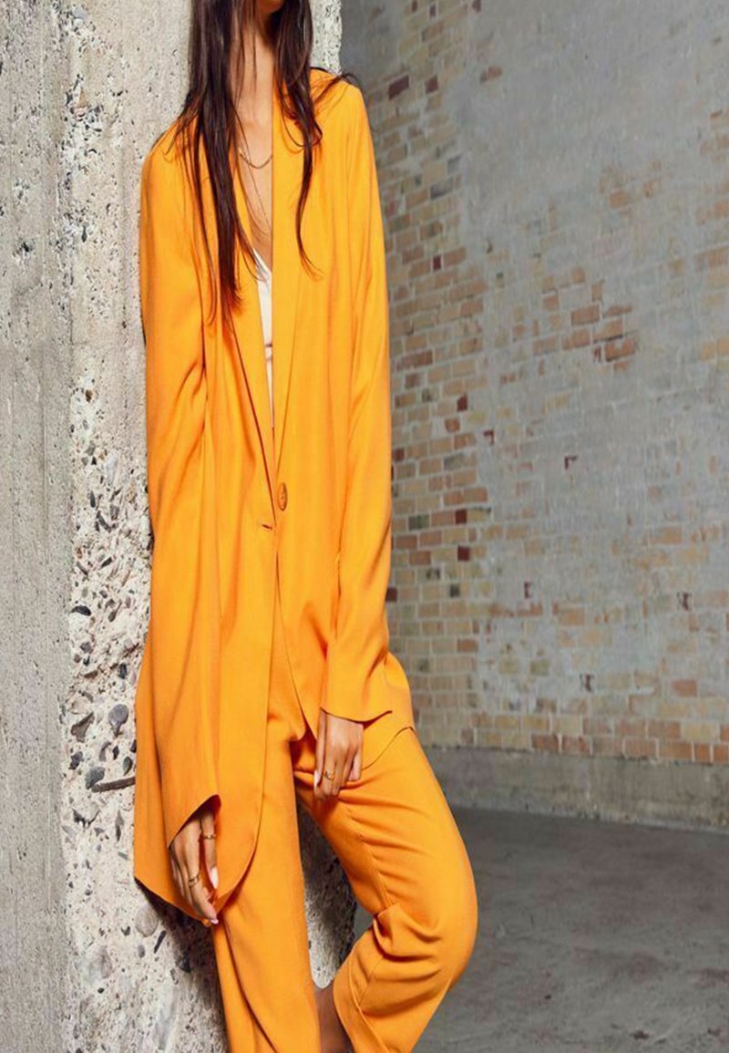 Femme portant un costume ample orange vif appuyée contre un mur en béton texturé avec un fond en briques apparentes.