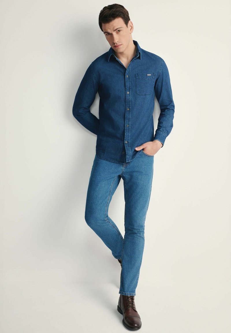 Granatowa koszula dżinsowa z kieszenią na piersi; zestawiona z obcisłymi niebieskimi dżinsami i brązowymi skórzanymi butami; posiada kołnierzyk zapinany na guziki i długie rękawy.