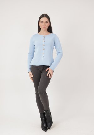 Donna in piedi che indossa un top a maniche lunghe azzurro con bottoni d'oro, jeans skinny scuri e stivaletti neri con punta appuntita.