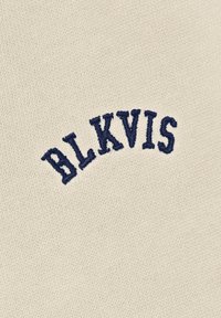 Granatowy wyhaftowany napis "BLKVIS" w zakrzywionym kształcie na beżowym, dzianinowym tle.