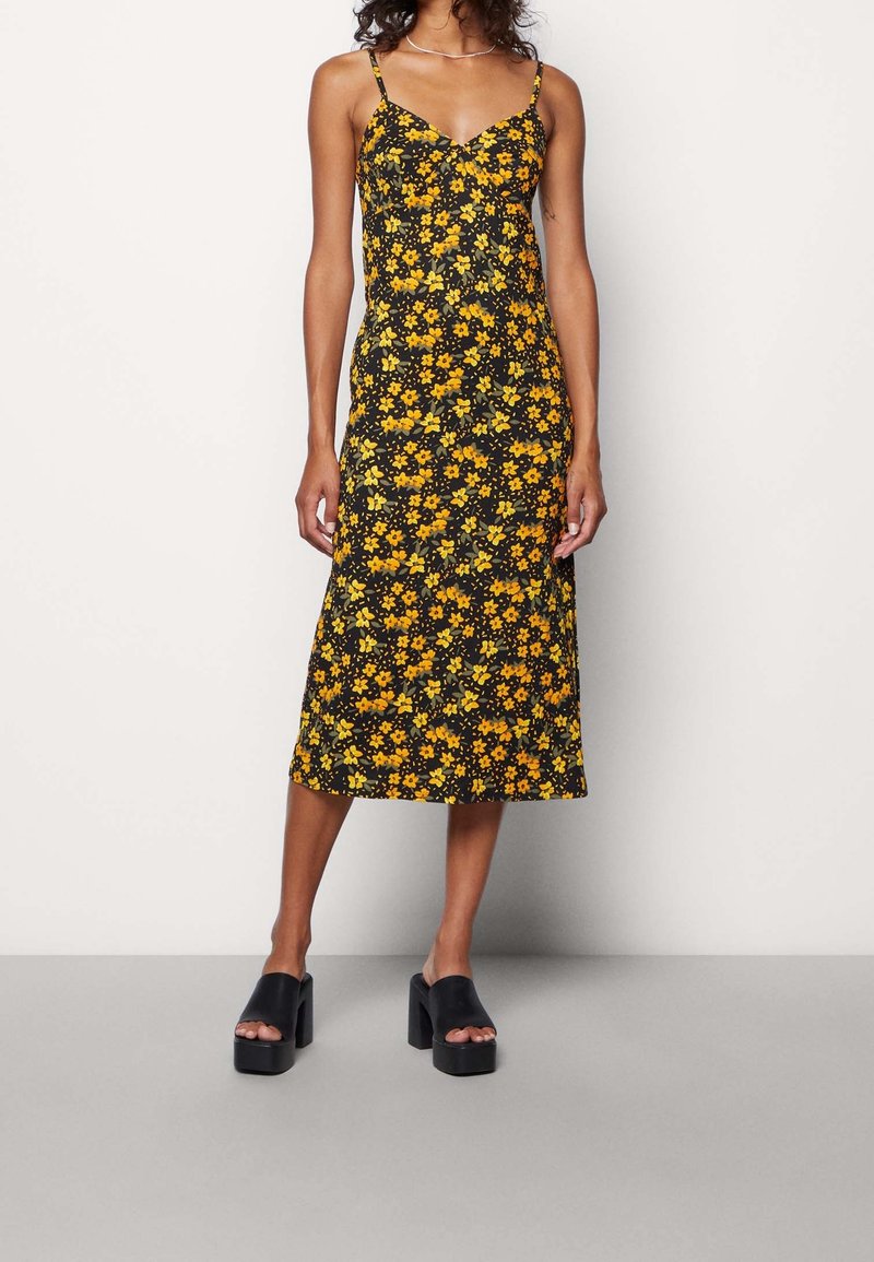 Svart blommig midiklänning med gula blommor, tunna axelremmar och en figursydd överdel. Kombineras med kraftiga svarta platåsandaler.