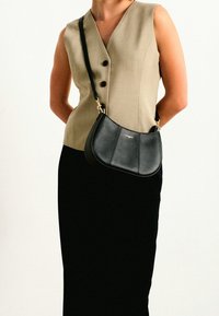 Borsa in pelle nera con una forma curva e hardware dorato, indossata a tracolla. Il modello indossa un gilet beige e una gonna nera.