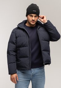 Granatowa kurtka puchowa z pikowanym wzorem, zamkiem błyskawicznym i kapturem, zestawiona z ciemnym beanie. Model ma na sobie granatowy sweter i jasnoniebieskie dżinsy.