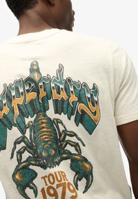 Cream-colored t-shirt featuring a large green and gold scorpion graphic with "Superdry" and "Tour 1979" text below, in distressed print.