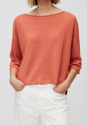 Femme portant un pull orange ample à manches longues avec un pantalon blanc, mains dans les poches, cadrée aux épaules et aux hanches.