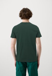 Persona con cabello corto y rizado castaño, vistiendo una camiseta verde oscuro y pantalones cargo azul verdoso, de espaldas contra un fondo blanco liso.