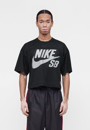 Jeune homme atteint de vitiligo portant un t-shirt en mesh noir Nike SB et un pantalon noir avec des bandes rouges, debout devant un fond blanc uni.