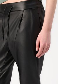 Sorte faux skinnbukser med en glatt tekstur, med folder og en elastisk snøring i livet. Synlig hånd hviler på buksebenet.