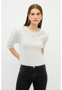 Weißer Strickpullover mit kurzen Puffärmeln und dekorativen Perlenakzenten am Ausschnitt, kombiniert mit hochgeschnittenen schwarzen Jeans.